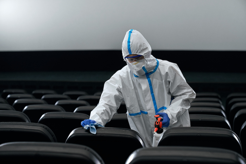 Man in white protective anti-virus suit wiping chairs in cinema. Concept of disinfection cinema hall.
